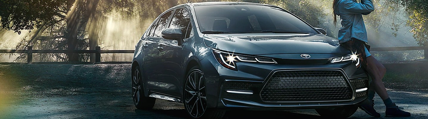 A dramatic 3/4 front view of a 2022 Toyota Corolla parked in the woods with a young woman leaning against the driver side.