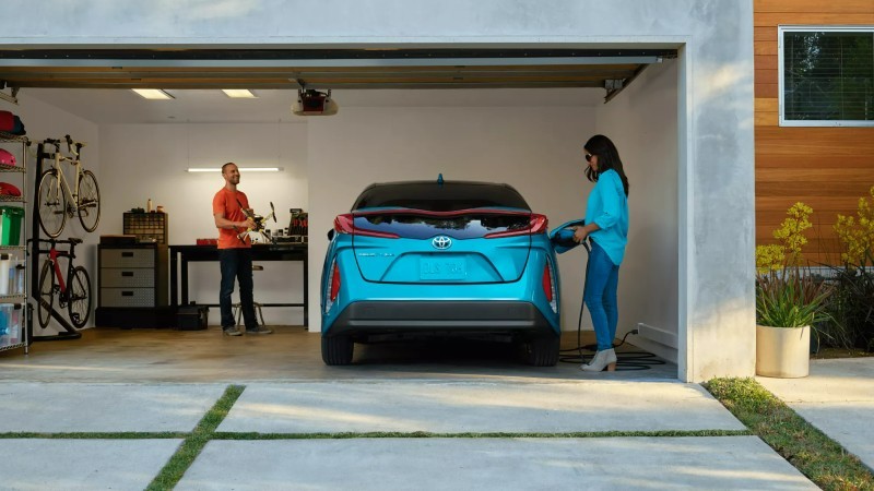 A shot into a garage from the driveway, showing a 2022 Prius parked inside with a woman plugging in the charger.
