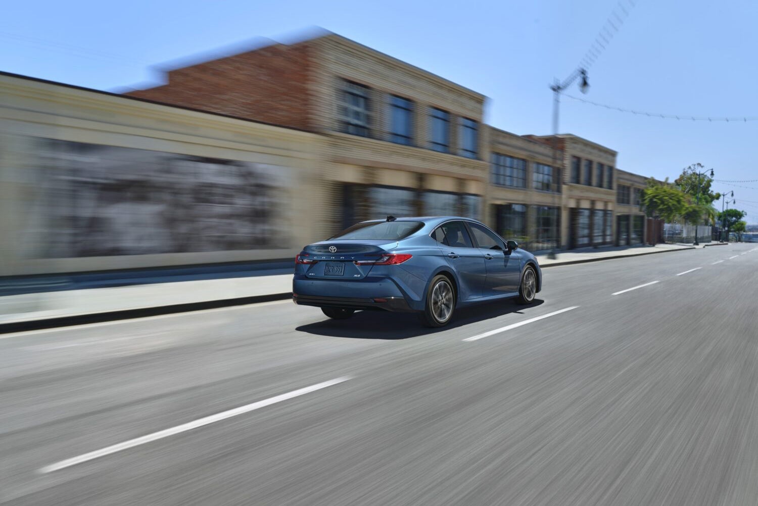A new model blue Toyota Camry sedan is shown from the rear three-quarter view, driving quickly down a wide, empty city street. Motion blur on the foreground and background highlights the car's speed. A row of brick and stucco commercial buildings are visible on the left.