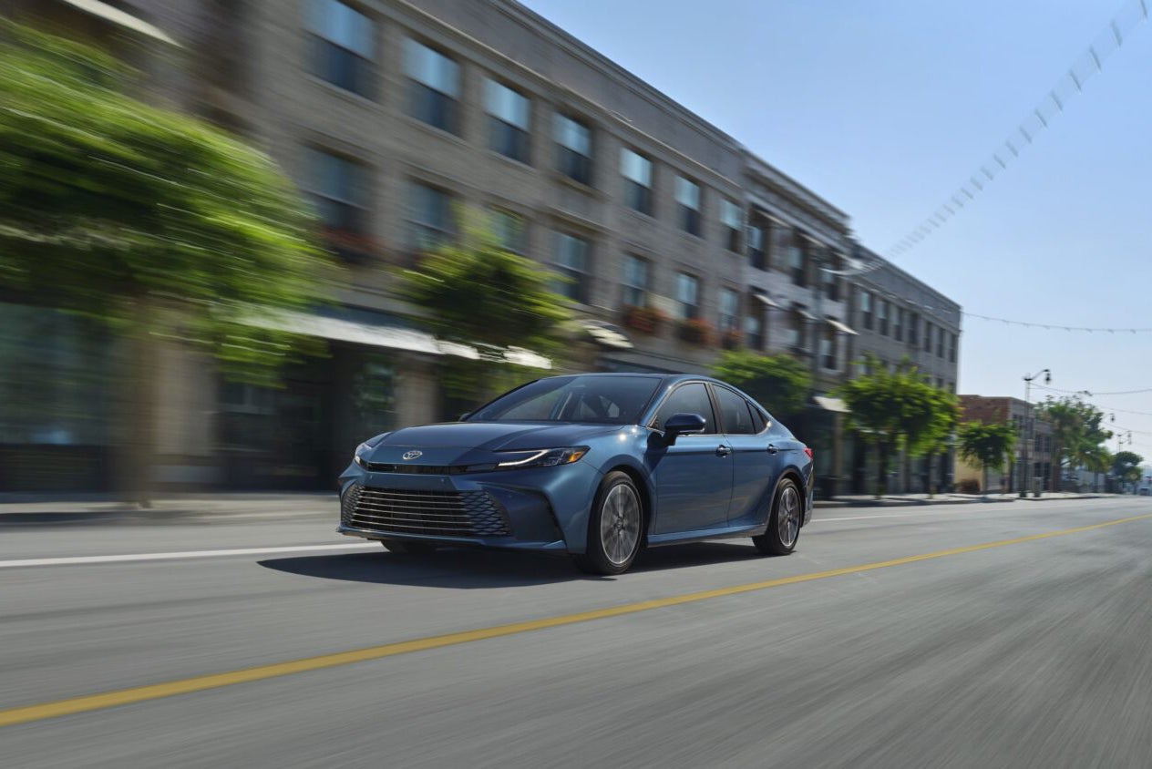 A new model blue Toyota Camry sedan drives on a street in a city, with motion blur indicating speed. A row of multistory residential or commercial buildings and trees line the right side of the street.
