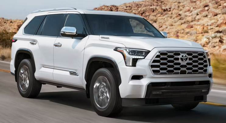A white Toyota Sequoia SUV drives on a paved road with rocky, dry hills in the background under daylight.