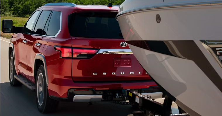 A red Toyota Sequoia SUV towing a boat on a trailer, driving on a road with green foliage.
