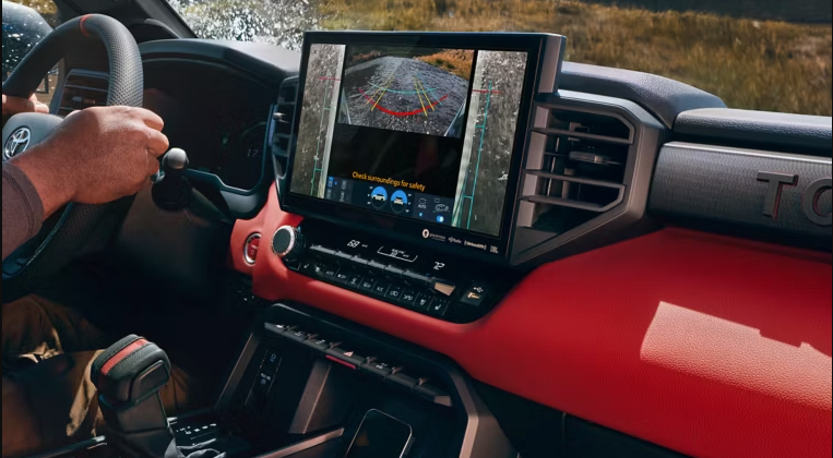 A car's interior shows a large touchscreen displaying a 360-degree camera view of an off-road path, with a driver's hand on the steering wheel and a red dashboard.