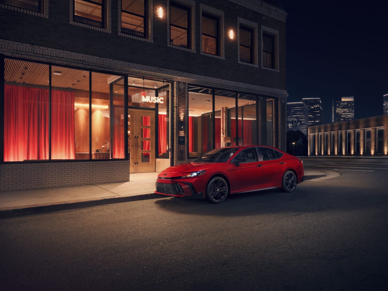 A red American made Toyota Camry in Phoenix is parked facing left by the curb of a city street next to a music venue at night.
