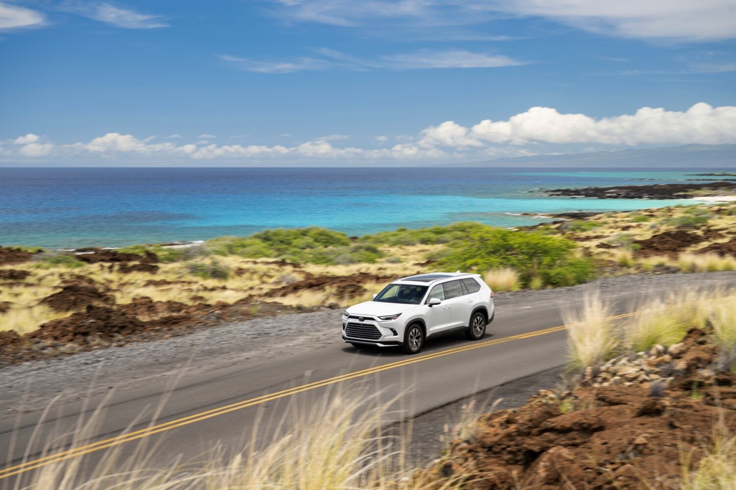 A white American made Toyota Grand Highlander in Phoenix drives towards the viewer to the left on a coastal road with a beach and dunes in the background, seen from a high angle.