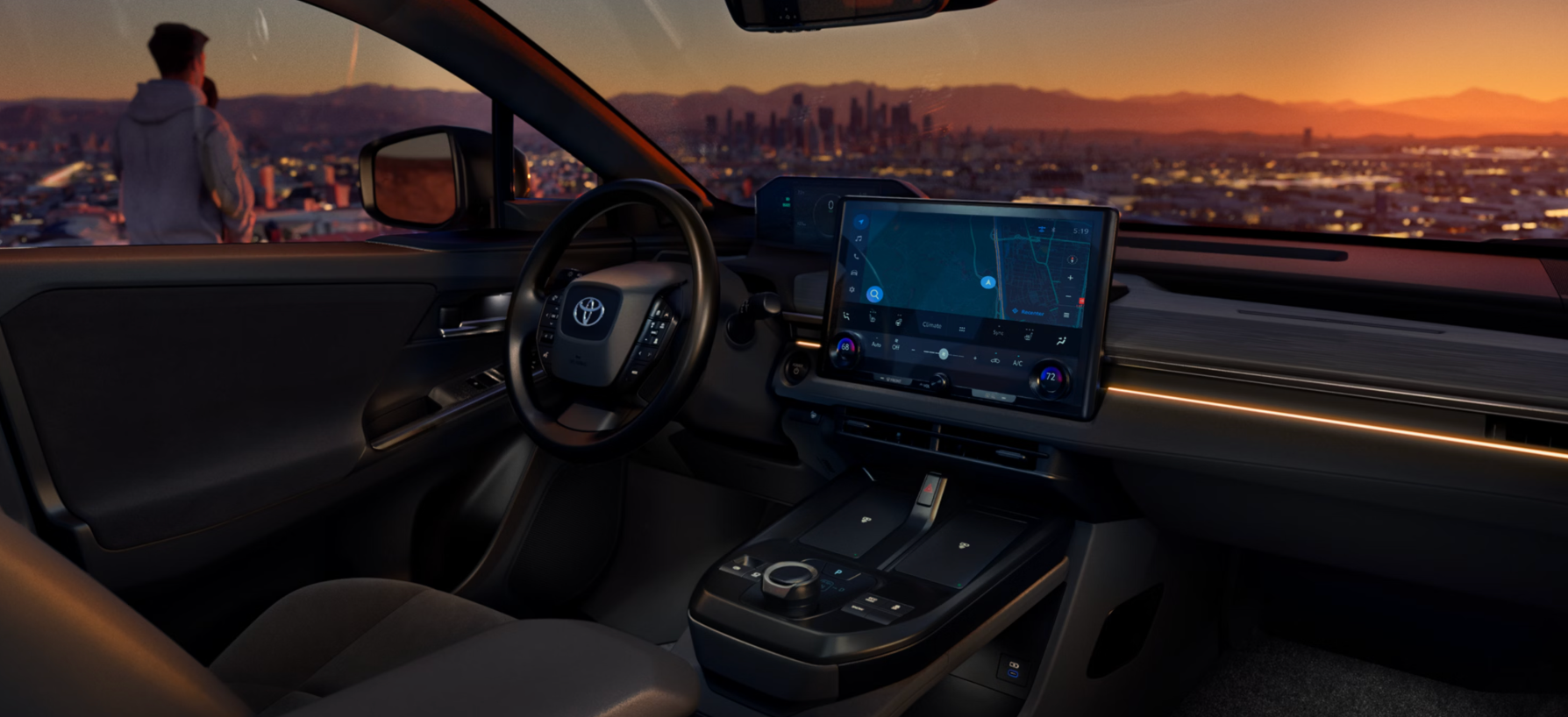 Interior shot of a dark-colored vehicle cabin, showing the dashboard, steering wheel, and large floating central touchscreen. The screen displays a navigation map. The vehicle is parked overlooking a vast, distant city skyline at sunset, casting warm orange and pink light. A person in a light-colored hoodie is visible standing outside the open driver's door, looking out at the view.