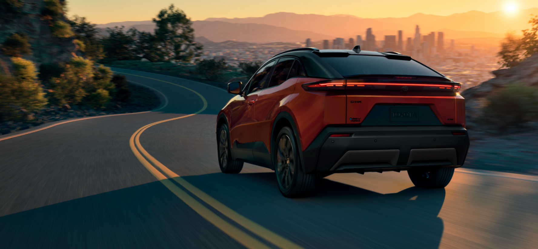 Rear three-quarter view of an orange or red compact crossover SUV driving on a winding, two-lane road at sunset. The vehicle features a black roof and a full-width LED taillight bar that is illuminated. In the distance, a large city skyline is visible against the warm orange glow of the setting sun over rolling hills.