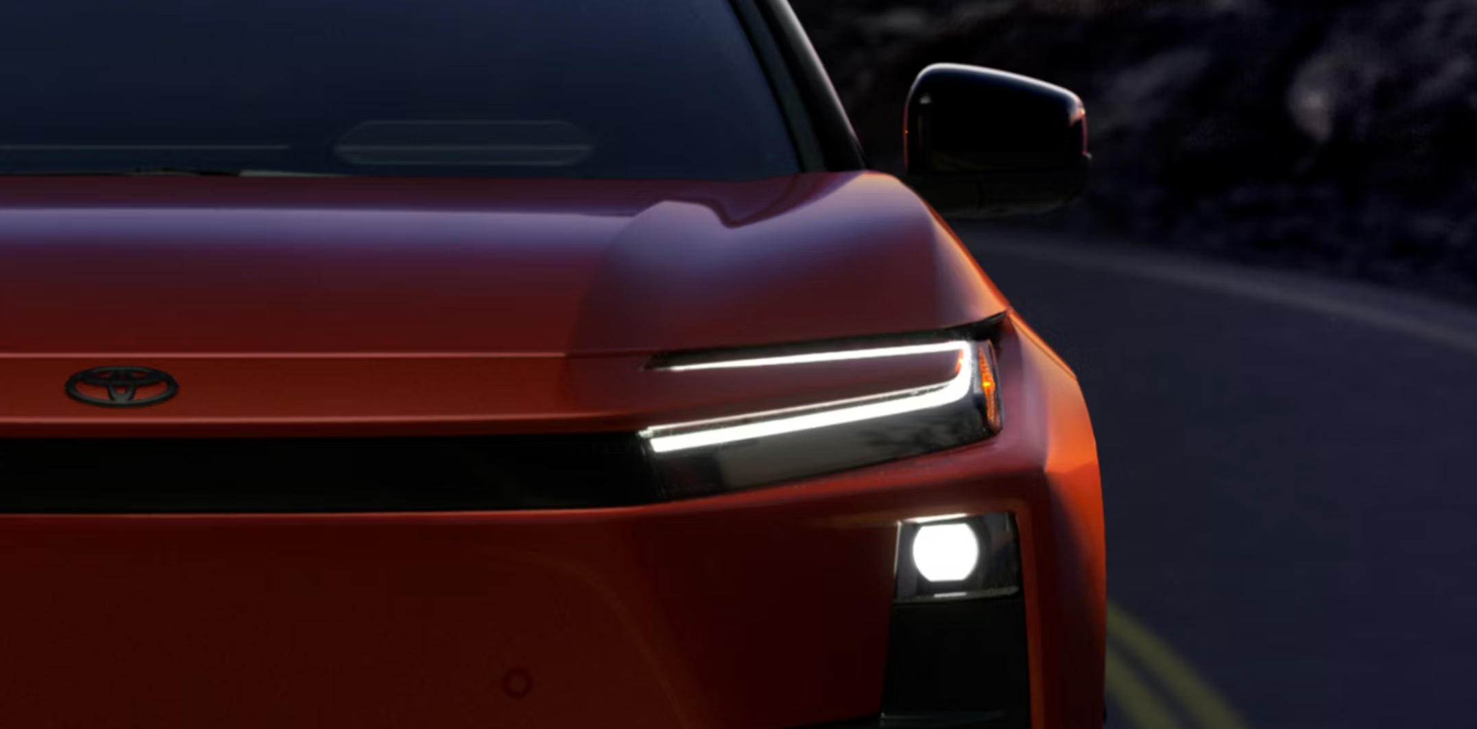 A close-up, low-light shot of the front of a burnt orange or red vehicle, focusing on the driver's side headlight assembly. The light features a distinctive white, horizontal C-shaped LED daytime running light and a lower circular projector fog light. A dark road and blurred mountain-like background are visible in the distance.