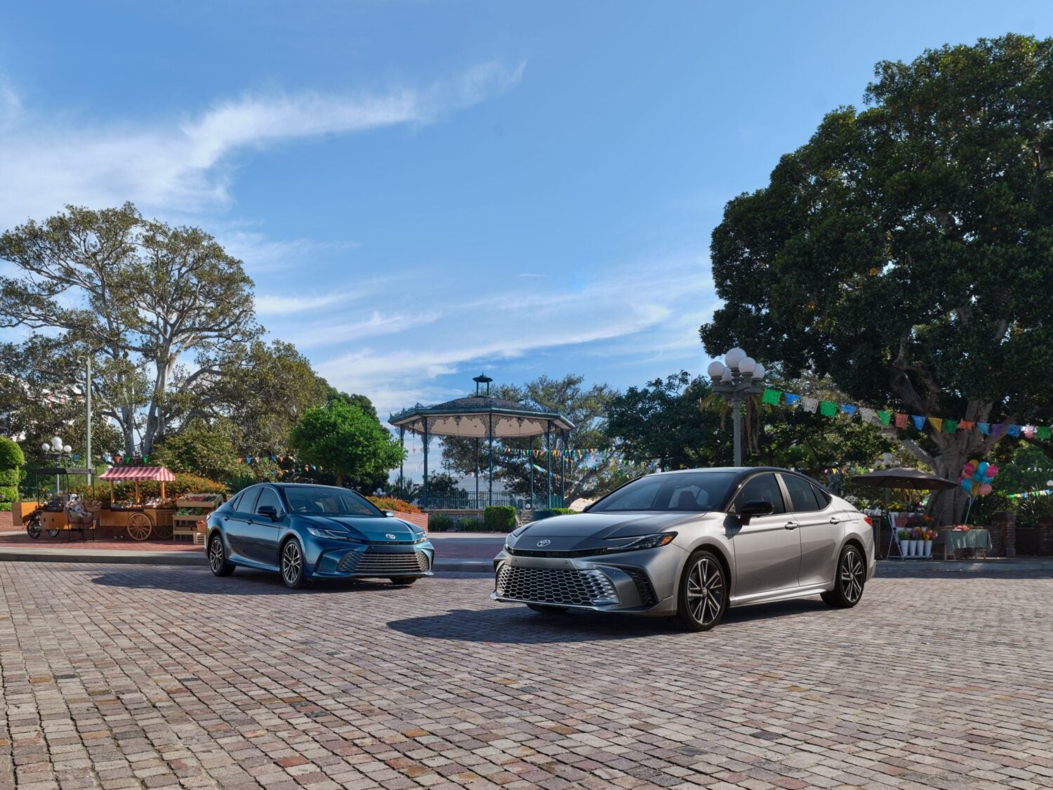 Two 2026 Toyota Camrys in Phoenix (blue, left/back; silver, right/front) are parked angled inwards facing the viewer on a cobblestone patio with a city park in the background.
