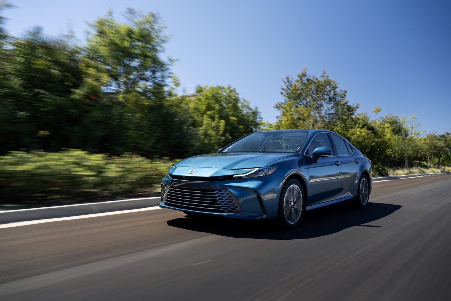 A blue 2026 Toyota Camry in Phoenix drives towards the viewer to the left on a two lane road with trees in the background.