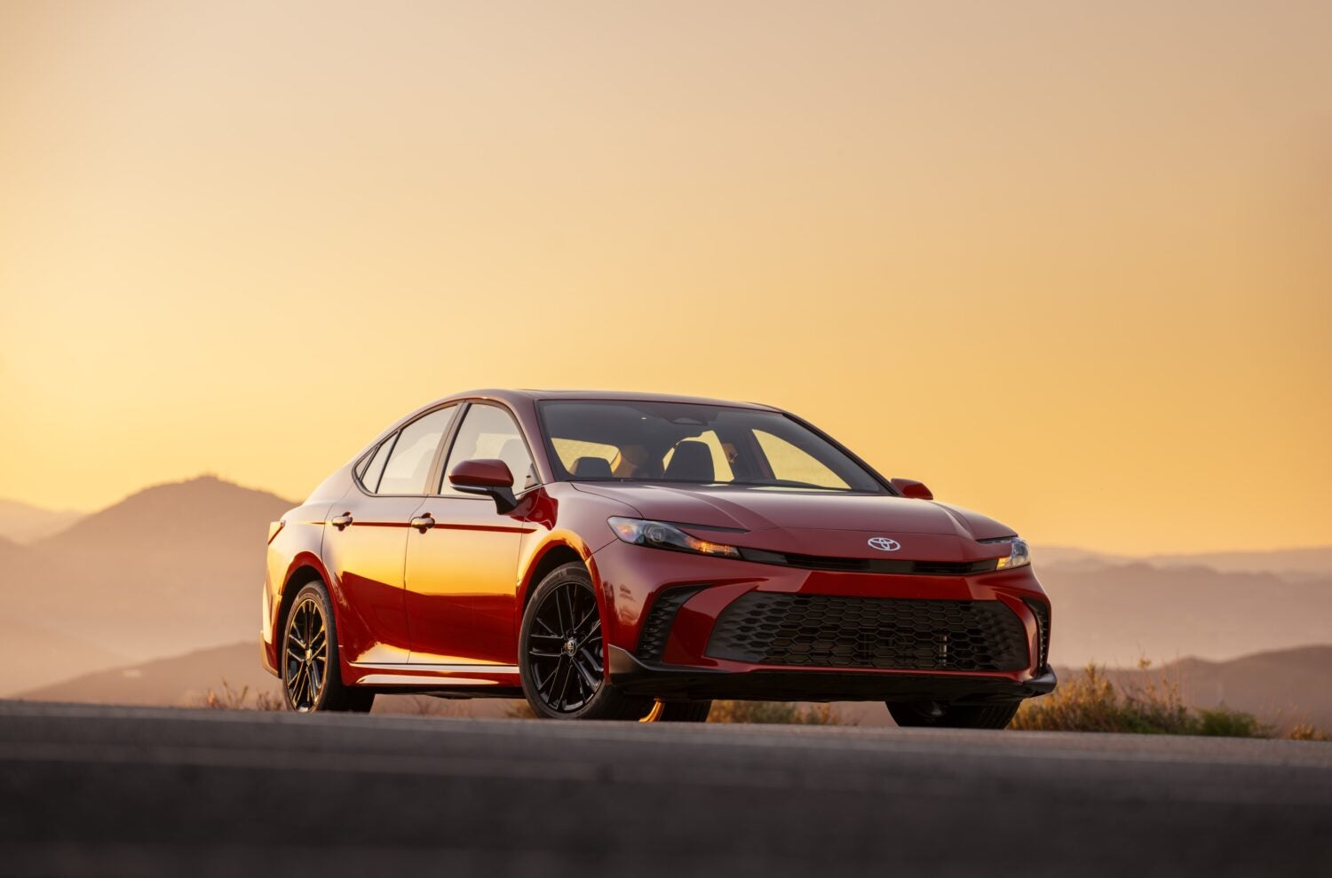 A red new Toyota Camry in Phoenix is parked facing the viewer to the right seen from a low angle on a desert highway with the orange light of a sunset and faded mountains in the background.