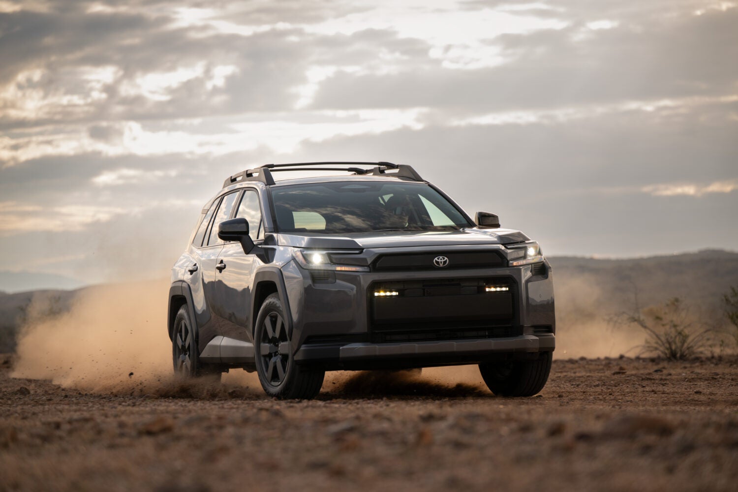 A grey 2026 Toyota Rav4 Woodland in Phoenix drives towards the viewer to the right on a dirt trail kicking up dust at sunset.